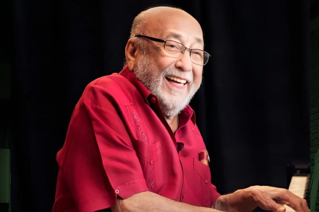 El legendario pianista Eddie Palmieri, conocido como 'El Sol de la Música Latina', sonríe sentado al piano. Su fallecimiento es lamentado por figuras como Willie Colón, dejando un gran legado en la salsa y el latin jazz.