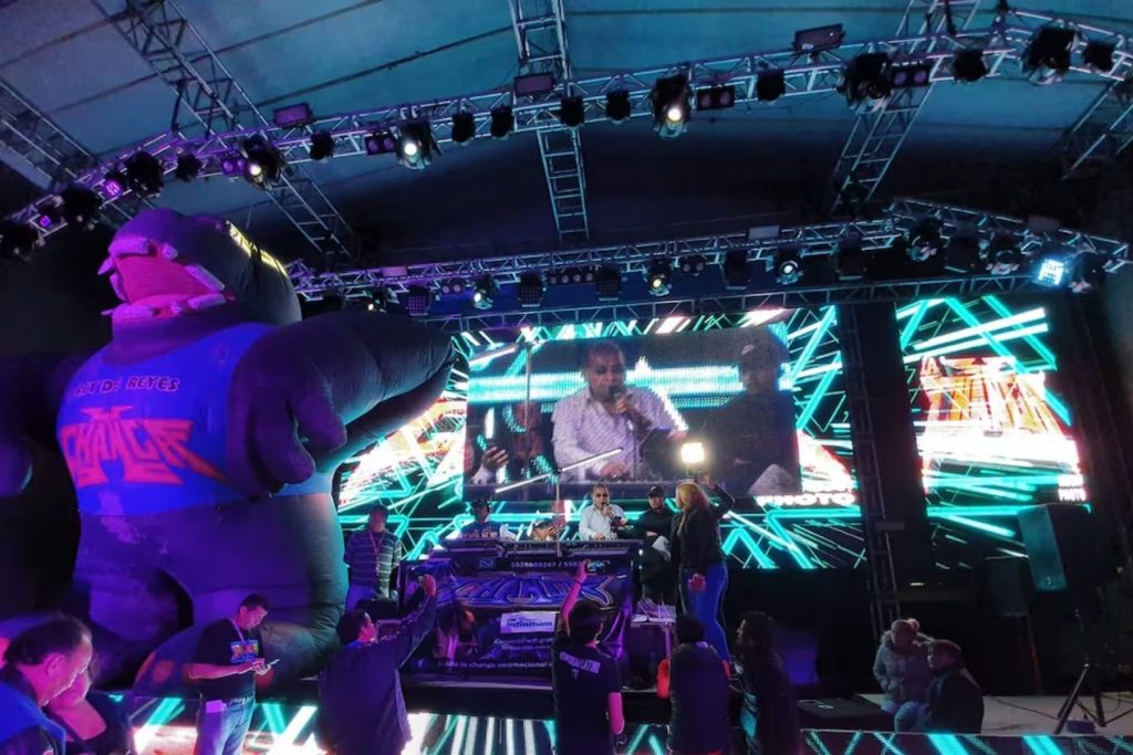 El ambiente de un baile con Sonido La Changa por la noche, con el escenario iluminado por luces de neón y la pantalla gigante mostrando a Ramón Rojo en acción. La imagen captura la energía y la fiesta de la cultura sonidera, un pilar del entretenimiento en colonias populares.