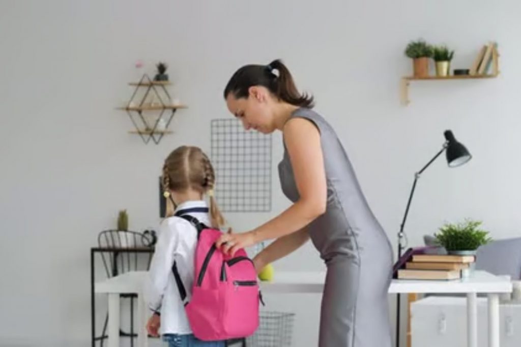 Una madre ayuda a su hija a ponerse la mochila, listos para salir a la escuela. La nota explora los desafíos de la rutina matutina en el regreso a clases.