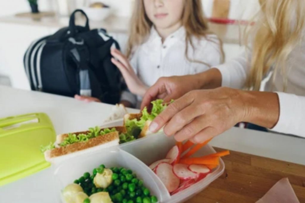 Una madre preparando un lunch saludable para su hija antes de ir a la escuela. La nota ofrece consejos para organizar la rutina matutina y evitar el estrés.