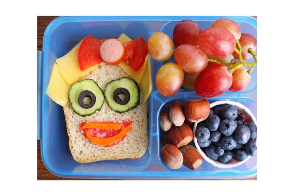 Un lunch escolar creativo con un sándwich decorado como una cara, frutas y nueces. La nota ofrece ideas fáciles y divertidas para el lunch de los niños.