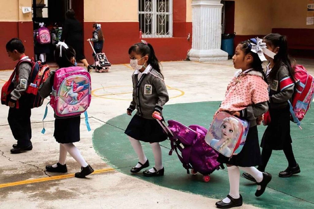 Niños de primaria con uniformes y mochilas en el patio de la escuela. La nota informa sobre el inicio del ciclo escolar 2025-2026 de la SEP.