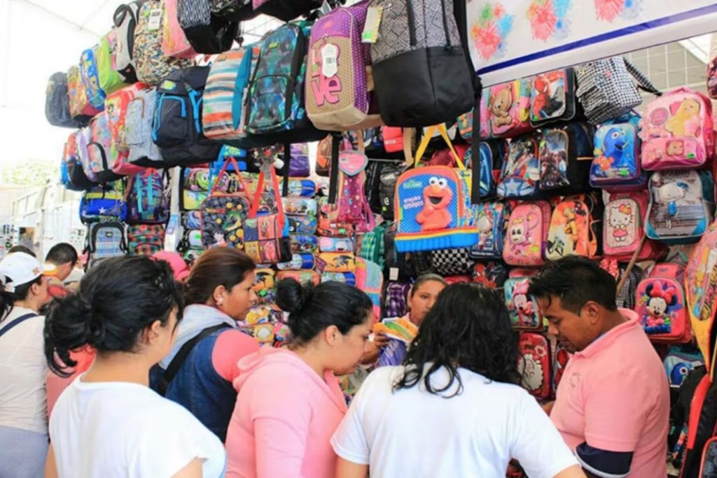 Padres de familia eligiendo mochilas en un puesto para el regreso a clases. La nota informa sobre los precios y la calidad de los útiles escolares según Profeco.