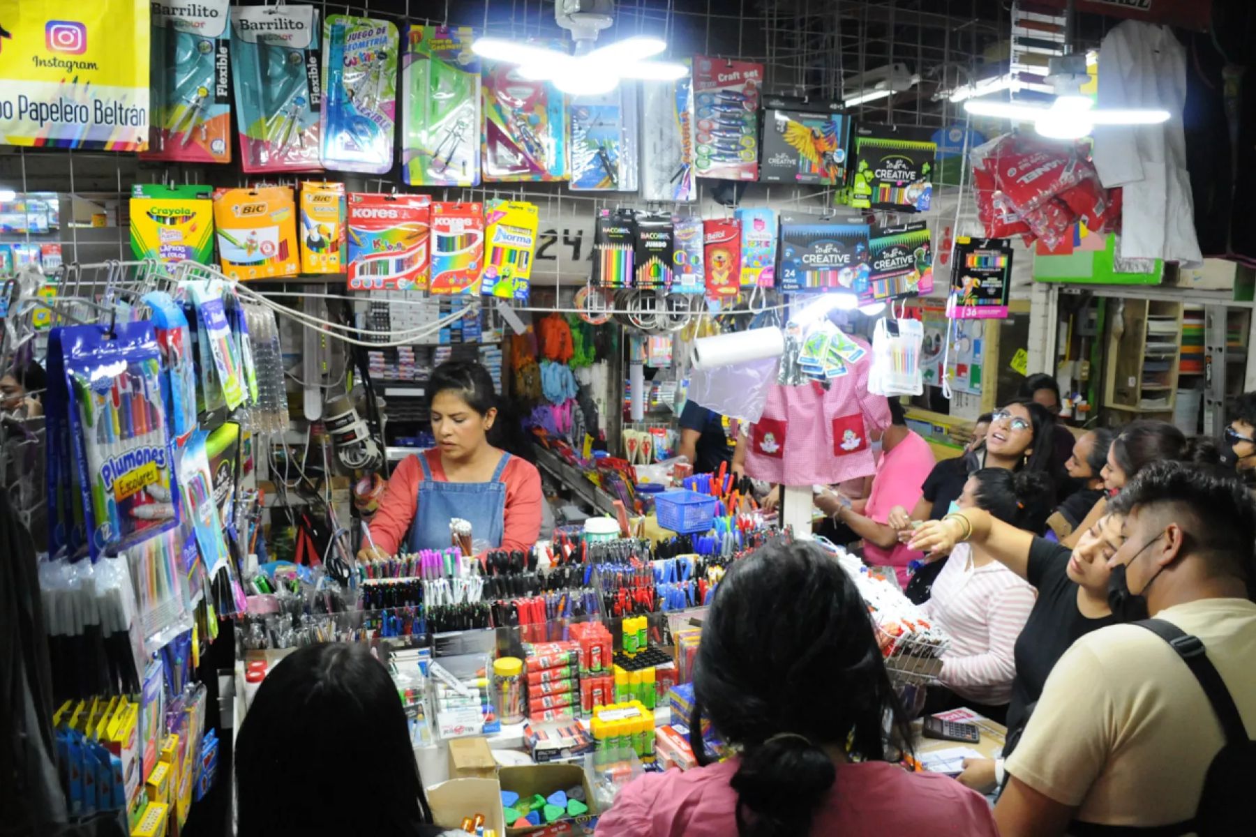 Un puesto de papelería atendiendo a muchos clientes por la noche. La nota da consejos para las compras de última hora para el regreso a clases.