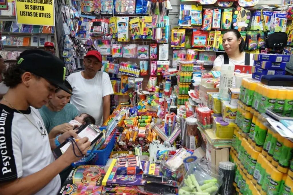 Una familia surtiendo la lista de útiles escolares en un puesto del centro. La nota informa sobre el costo promedio del regreso a clases para este ciclo escolar.