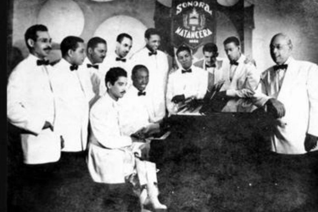 Los integrantes de la Sonora Matancera reunidos alrededor del piano. La nota explora el sonido único de la orquesta y el talento de sus músicos.
