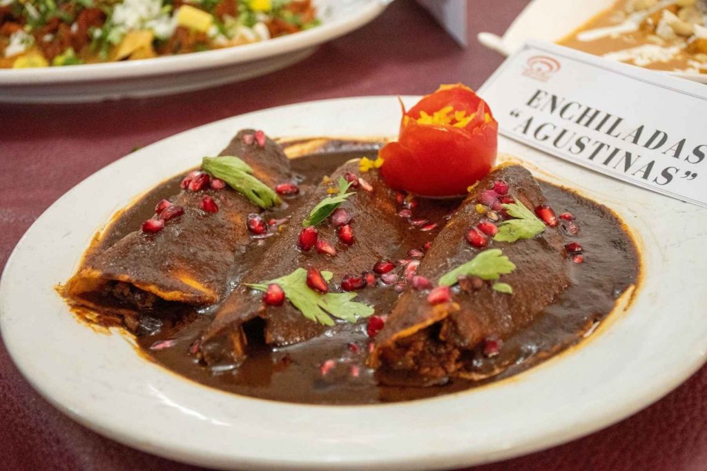 Un plato de enchiladas "Agustinas" con mole y granada. La nota informa sobre la Feria de la Enchilada en Iztapalapa y la variedad de platillos que se ofrecen.
