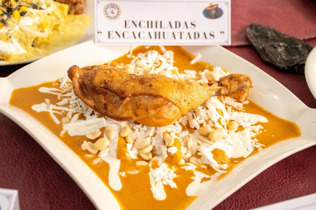 Un plato de enchiladas encacahuatadas con una pieza de pollo. La nota invita a la Feria de la Enchilada en Iztapalapa para disfrutar de este y otros platillos.