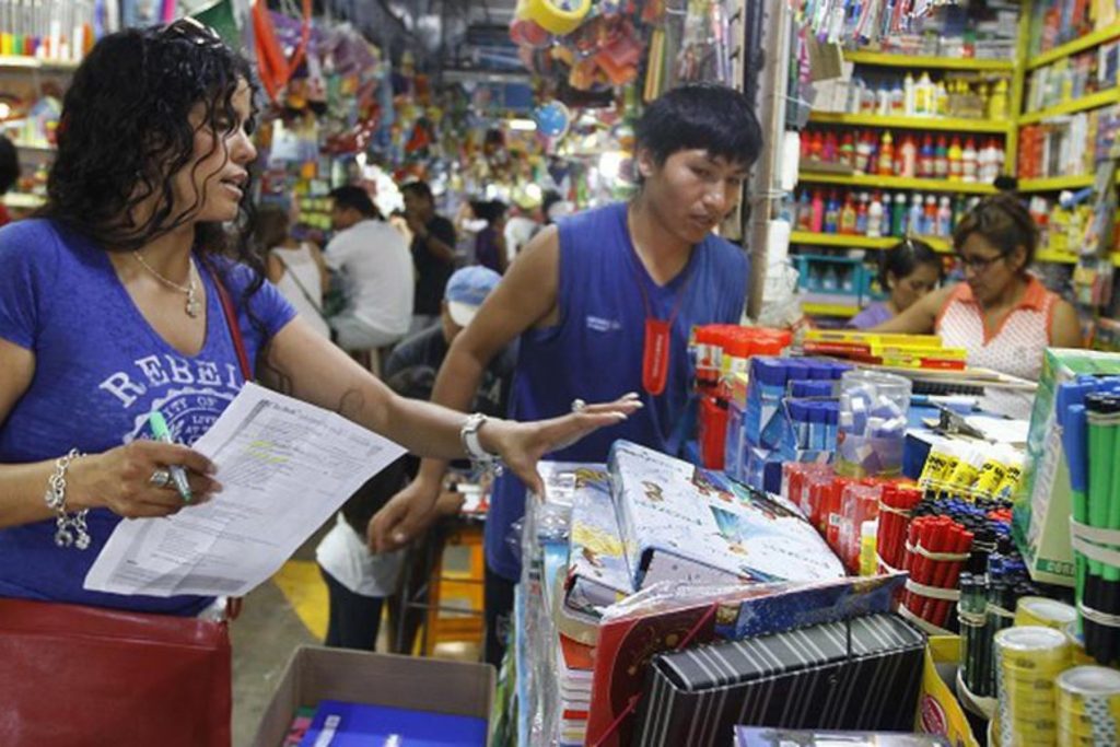 Una madre de familia con la lista en mano, pidiendo los útiles escolares a un vendedor. La nota ofrece una guía para no salirse del presupuesto en este regreso a clases.