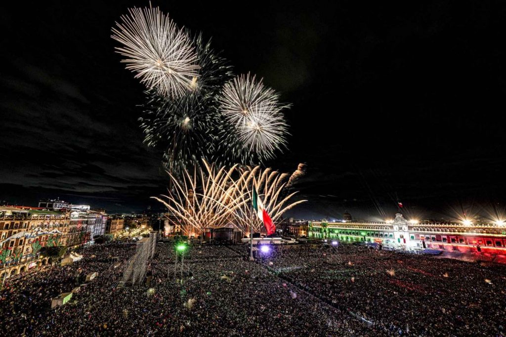 Vista panorámica del Zócalo de la Ciudad de México lleno de gente durante el Grito. La nota informa sobre los artistas que se presentarán en la celebración.