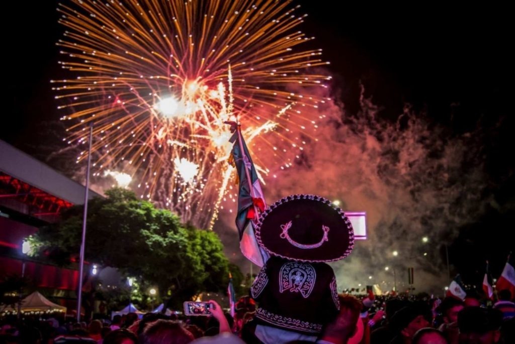 Una persona con sombrero de charro y una bandera de México viendo los fuegos artificiales. La nota celebra el orgullo y la alegría de las Fiestas Patrias.
