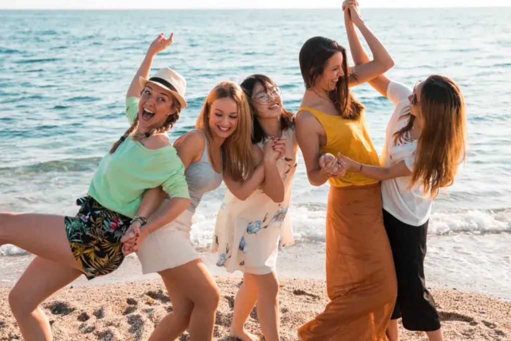 Un grupo de cinco amigas jugando y riendo en la playa. La nota habla sobre la importancia de crear recuerdos y vivir experiencias juntas para fortalecer la amistad.
