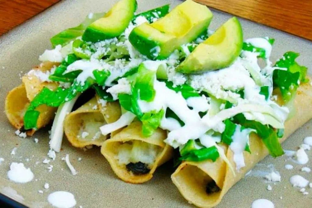 Un plato de flautas de papa con lechuga, crema, queso y aguacate. La nota ofrece recetas de antojitos económicos para celebrar el Grito.