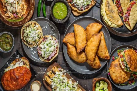 Una mesa llena de antojitos mexicanos como sopes, empanadas y tacos. La nota ofrece una guía completa para organizar tu cena del 15 de septiembre.