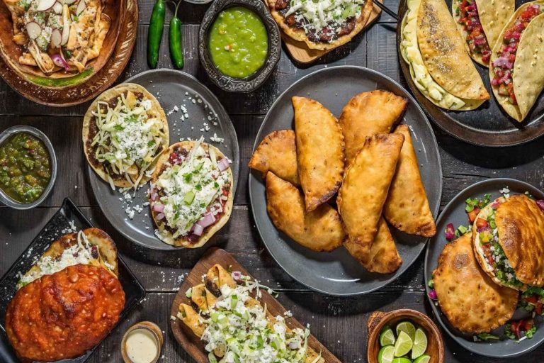 Una mesa llena de antojitos mexicanos como sopes, empanadas y tacos. La nota ofrece una guía completa para organizar tu cena del 15 de septiembre.