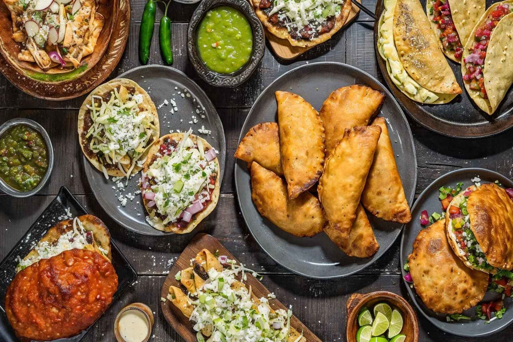 Una mesa llena de antojitos mexicanos como sopes, empanadas y tacos. La nota ofrece una guía completa para organizar tu cena del 15 de septiembre.
