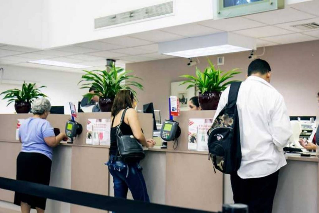 Personas formadas en el interior de un banco para ser atendidas en ventanilla. El artículo detalla que los bancos sí operarán el 15 de septiembre pero cerrarán el 16.