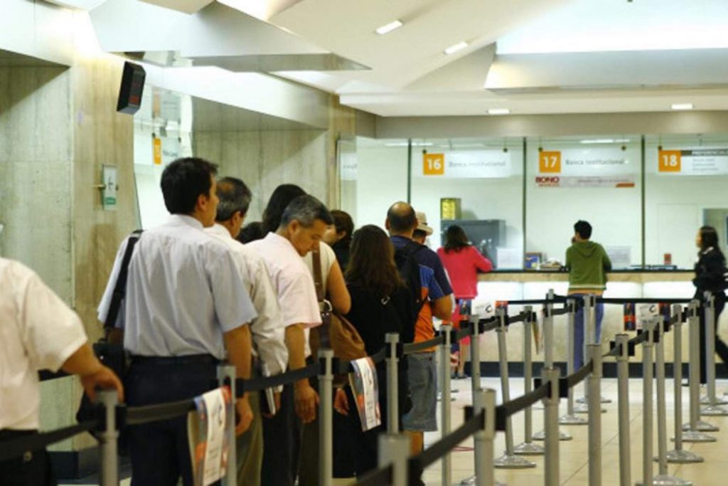 Una larga fila de personas esperando para realizar operaciones en un banco. La nota ofrece una guía sobre los horarios de los bancos en Fiestas Patrias.