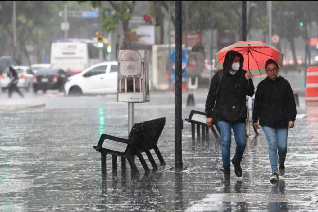 Dos mujeres caminando bajo la lluvia con un paraguas en una calle de la CDMX. El artículo detalla el pronóstico del clima para el 15 de septiembre.