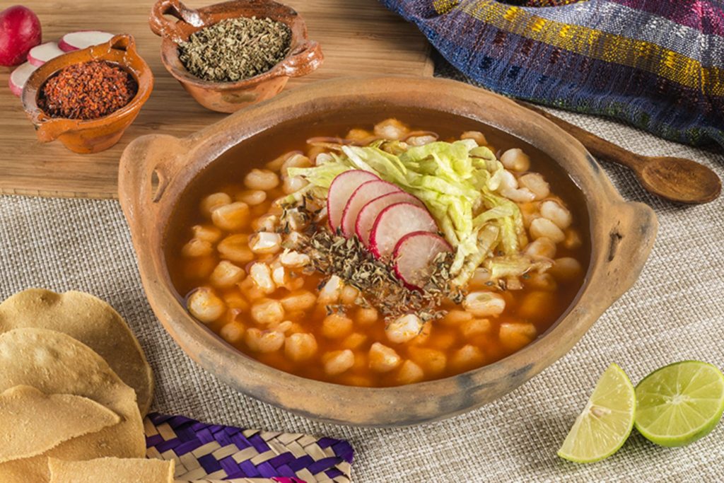 Un plato de pozole rojo servido en un tazón de barro con todas sus guarniciones. La nota ofrece la receta de la abuela para que te quede delicioso.