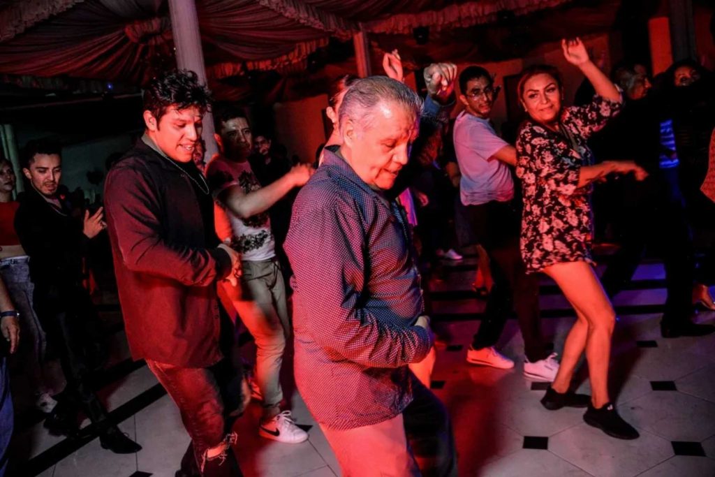Personas de todas las edades bailando cumbia en un salón de fiestas. La nota celebra la música y el baile que unen a la familia en las celebraciones.