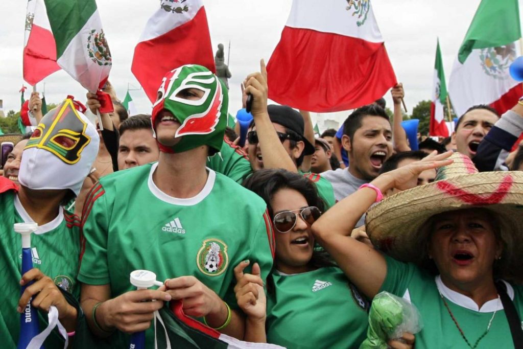 Aficionados mexicanos con playeras verdes y banderas celebrando un gol. La nota habla sobre la pasión de la afición de cara al Mundial 2026.