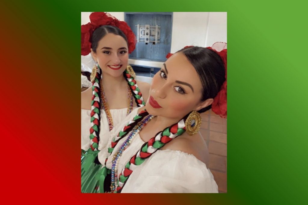 Dos mujeres con peinados de trenzas largas y flores rojas, vestidas para la Noche Mexicana. La nota ofrece ideas de peinados para celebrar el 15 de septiembre.